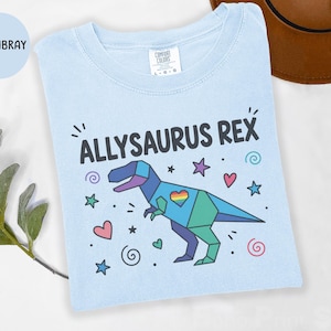 Puede incluir: Una camiseta azul claro con el texto "ALLYSAURUS REX" sobre un gráfico de dinosaurio geométrico. El dinosaurio está compuesto por segmentos azules, verdes y morados con un corazón arcoíris en el pecho, rodeado de pequeñas estrellas, corazones y espirales.