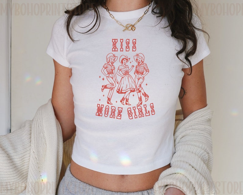 K&ouml;nnte beinhalten: Wei&szlig;es, bauchfreies T-Shirt mit einer roten Grafik von drei Frauen in Cowboyh&uuml;ten, die tanzen, mit dem Text "Kiss More Girls".