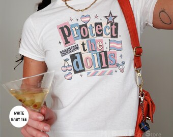 Könnte beinhalten: Ein weißes Baby-T-Shirt mit einem Grafikdruck, der den Text Protect the dolls in schwarzer, rosa und blauer Schrift zeigt. Das Design enthält Illustrationen eines Herzens, eines Sterns, einer Flagge, Kirschen und eines Geschlechtersymbols.
