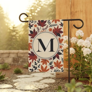 May include: A fall-themed garden flag with a cream background and a large, black letter "M" in a circle. The flag is decorated with orange, red, and brown leaves.