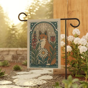 May include: A garden flag with a decorative illustration of a fox amidst flowers and a snowflake. The fox is depicted in brown and white, with a calm expression. The flowers include red foxgloves, a white daisy, and a red tulip. The flag is displayed outdoors.