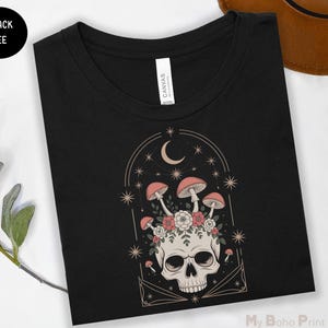 May include: Black tee shirt featuring a skull with mushrooms, flowers, and a crescent moon design. The graphic is in shades of pink, beige, and white. The shirt has a crew neck and a tag that says "CANVAS". The words "BLACK TEE" are in a black circle.
