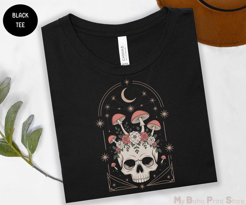 May include: Black tee shirt featuring a skull with mushrooms, flowers, and a crescent moon design. The graphic is in shades of pink, beige, and white. The shirt has a crew neck and a tag that says "CANVAS". The words "BLACK TEE" are in a black circle.