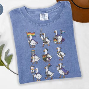 Pode incluir: Uma t-shirt de cor azul ganga com uma impressão gráfica de nove gansos brancos vestidos como cavaleiros e guerreiros medievais. Os gansos são retratados com vários acessórios, incluindo espadas, escudos, capas e bandeiras com cores arco-íris.