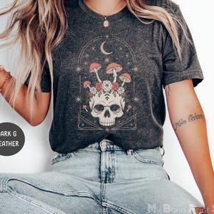 May include: Dark heather gray t-shirt featuring a skull design with mushrooms, flowers, and a crescent moon. The graphic is in shades of red, pink, and white. The shirt has a crew neck and short sleeves.
