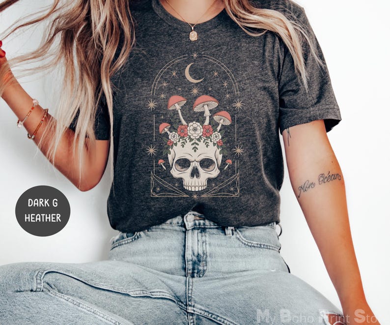 May include: Dark heather gray t-shirt featuring a skull design with mushrooms, flowers, and a crescent moon. The graphic is in shades of red, pink, and white. The shirt has a crew neck and short sleeves.