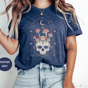 May include: Heather navy blue t-shirt featuring a skull design with mushrooms, flowers, and a crescent moon. The graphic is in a vintage style with a celestial theme. The shirt is made of a soft material.