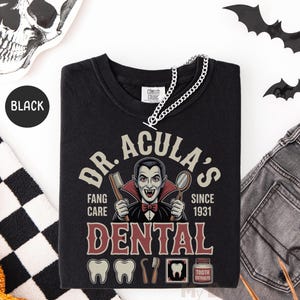 May include: Black crew neck t-shirt with a graphic that reads "DR. ACULA'S DENTAL" featuring a vampire illustration. The design includes dental tools and the text "FANG CARE SINCE 1931". A silver chain necklace is draped over the shirt.