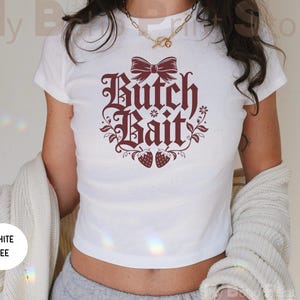 May include: A white short-sleeved t-shirt featuring a dark red graphic print on the chest. The design includes a bow at the top, the text "Butch Bait" in a stylized font, floral accents, and two strawberries at the bottom of the graphic.