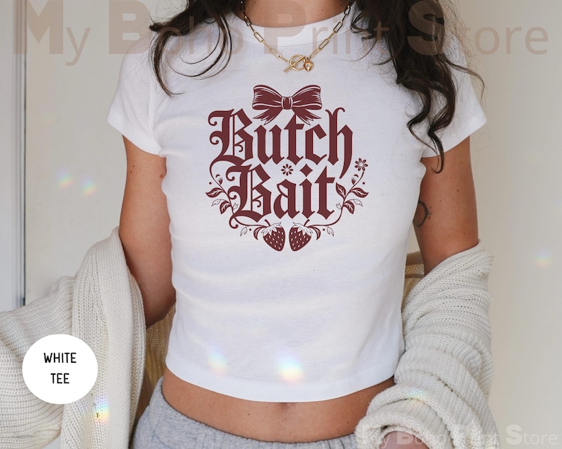 May include: A white short-sleeved t-shirt featuring a dark red graphic print on the chest. The design includes a bow at the top, the text "Butch Bait" in a stylized font, floral accents, and two strawberries at the bottom of the graphic.