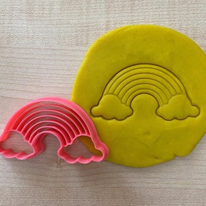 Può includere: Tagliapasta a forma di arcobaleno in plastica rosa con nuvole su entrambi i lati. Il tagliapasta è mostrato sopra l'impasto per biscotti giallo.