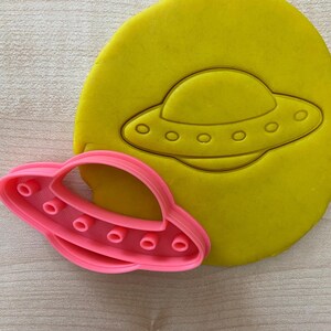 May include: A pink plastic UFO cookie cutter on a yellow background. The cookie cutter is used to cut out the shape of a UFO from cookie dough.