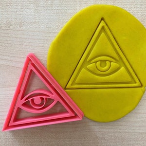 May include: A pink plastic cookie cutter shaped like a triangle with an eye inside. The cookie cutter is on a yellow surface.