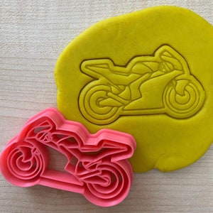 Peut inclure: Un emporte-pièce en plastique rose en forme de moto. L'emporte-pièce est présenté sur une pâte à modeler jaune.