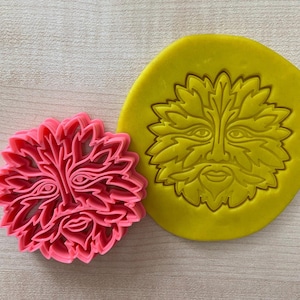 May include: Two cookie cutters, one pink and one yellow, shaped like a green man's face with leaves for hair.