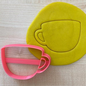 May include: A pink plastic cookie cutter shaped like a coffee cup. The cutter is on a yellow surface with a cutout of a coffee cup.