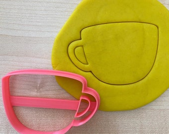 May include: A pink plastic cookie cutter shaped like a coffee cup. The cutter is on a yellow surface with a cutout of a coffee cup.