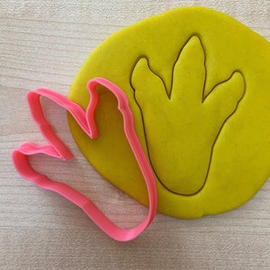 May include: A pink plastic dinosaur footprint cookie cutter on a yellow background.
