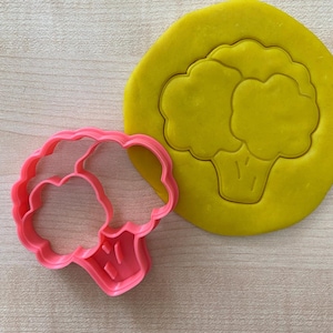 May include: A pink plastic cookie cutter shaped like a broccoli floret. The cutter is on a wooden surface next to a yellow piece of modelling clay with a broccoli impression.
