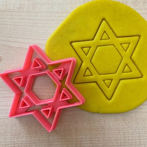 Op de afbeelding: Een roze plastic koekjesvormpje in de vorm van een zespuntige ster, ook wel bekend als de Ster van David, met een kleinere zespuntige ster erin. Het koekjesvormpje ligt op een houten oppervlak naast een stuk geel deeg met dezelfde vorm erop gedrukt.