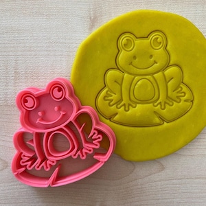 Peut inclure: Emporte-pièce en plastique rose en forme de grenouille assise sur un nénuphar. L'emporte-pièce est présenté à côté d'une empreinte jaune de l'emporte-pièce dans de la pâte à modeler.