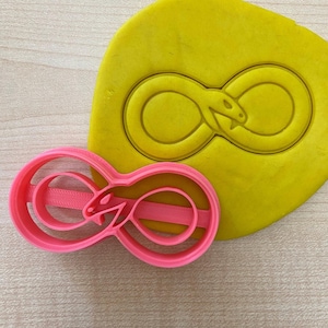 May include: Pink plastic cookie cutter shaped like an infinity symbol with a snake wrapped around it. The cutter is on a yellow surface.