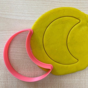 Könnte beinhalten: Ein rosa Mondförmiger Ausstecher neben einem gelben Teigstück mit einem Mondabdruck. Der Ausstecher ist aus Kunststoff und befindet sich links vom Teig, wobei er diesen teilweise überlappt.