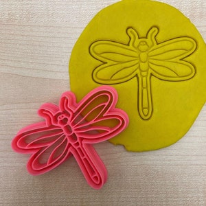Pode incluir: Um cortador de biscoitos de libélula de plástico rosa com um cortador de biscoitos de libélula de plástico amarelo no fundo.