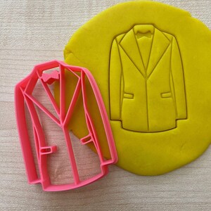 Peut inclure: Emporte-pièce en plastique rose en forme de veste de costume. L'emporte-pièce est sur un fond jaune.