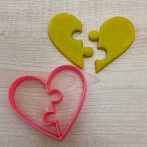 Puede incluir: Dos cortadores de galletas en forma de corazón, uno rosa y otro amarillo, se muestran sobre una superficie de madera. Ambos cortadores están diseñados con una forma de pieza de rompecabezas, creando un par a juego.