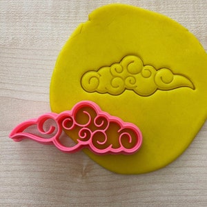May include: A pink plastic cookie cutter shaped like a cloud with swirls. The cutter is on top of a yellow piece of fondant.