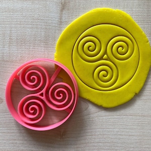 Può includere: Un tagliapasta in plastica rosa a forma di triskel, un simbolo celtico con tre spirali intrecciate. Il tagliapasta è su una superficie di legno accanto a un pezzo di pasta gialla con lo stesso disegno impresso su di esso.