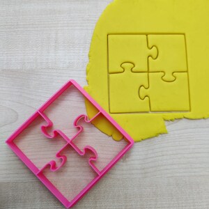 Pode incluir: Um cortador de biscoitos de plástico rosa em forma de quebra-cabeça de quatro peças. O cortador está em cima de uma massa amarela, também em forma de quebra-cabeça de quatro peças.
