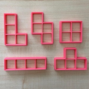 以下が含まれることがあります： テトリスのブロックの形をしたピンクのプラスチック製のクッキーカッターが5つ。ブロックは薄い茶色の木製の表面に一列に並んでいます。