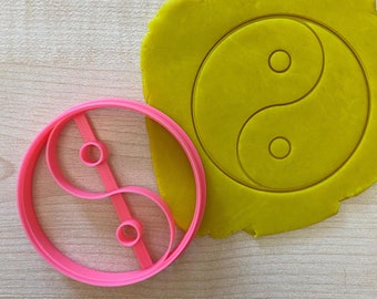 Może przedstawiać: Różowa plastikowa foremka do ciasteczek w kształcie symbolu yin yang. Foremka leży na żółtej powierzchni z wzorem yin yang.