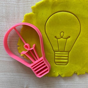 Könnte beinhalten: Ein rosa-farbener Plastik-Ausstecher in Form einer Glühbirne auf einer gelben Oberfläche. Der Ausstecher wird verwendet, um Glühbirnenformen in Teig zu erstellen.