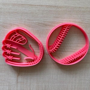 Puede incluir: Dos cortadores de galletas de plástico rosa, uno con forma de pelota de béisbol y el otro con forma de guante de béisbol.