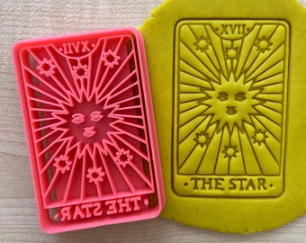 May include: A pink plastic cookie cutter shaped like a tarot card with the image of a sun with a face and the text "THE STAR" and "XVII" on it. The cookie cutter is on a yellow surface.