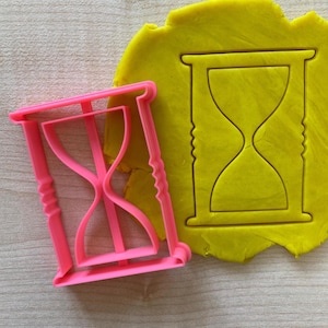 May include: Pink plastic hourglass cookie cutter with a yellow dough imprint of the same shape.