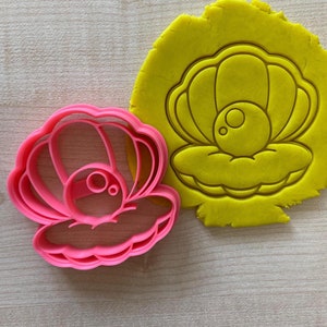 Op de afbeelding: Roze plastic koekjesvormpje in de vorm van een schelp met een parel erin. De vormpje ligt op een gele ondergrond.