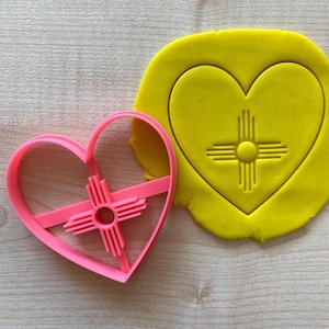 May include: Pink heart-shaped cookie cutter with a Zia symbol inside. The cutter is next to a yellow heart-shaped piece of cookie dough with the Zia symbol imprinted on it.