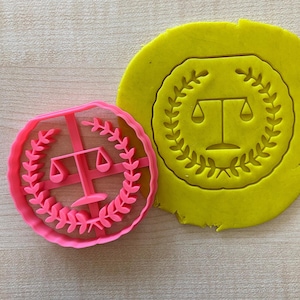 Könnte beinhalten: Rosaer Plastik-Ausstecher in Form einer Justitia-Waage mit einem Lorbeerkranz darum. Der Ausstecher liegt auf einer gelben Oberfläche.