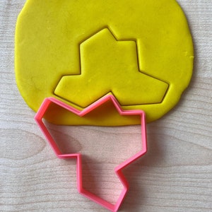 May include: A pink plastic cookie cutter shaped like a geometric star. The cutter is on a wooden surface with a yellow piece of modelling clay behind it.