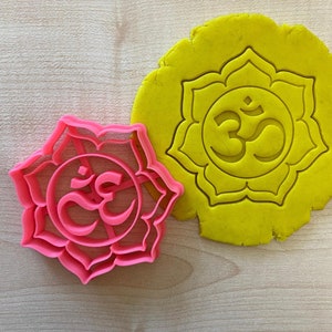 Può includere: Un tagliabiscotti in plastica rosa a forma di fiore di loto con il simbolo Om al centro. Accanto, un impasto giallo con il simbolo Om impresso. Il tagliabiscotti e l'impasto sono su una superficie di legno chiaro.