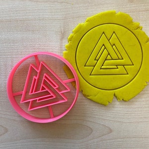 Può includere: Un tagliapasta di plastica rosa a forma di cerchio con un design a triangolo triplo all'interno. Il tagliapasta è accanto a un cerchio di pasta giallo con lo stesso design impresso su di esso.