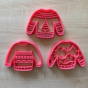 May include: Three pink plastic Christmas sweater cookie cutters. The cutters feature different designs, including a Christmas tree, snowflakes, and lights.