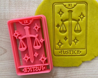 May include: Pink plastic cookie cutter shaped like a tarot card with the Justice card design. The card features a sword and scales with stars in the background. The text "JUSTICE" and Roman numeral "XI" are on the card.