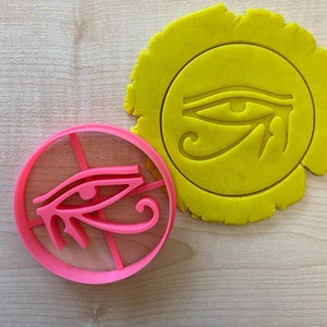 Op de afbeelding: Een roze plastic koekjesvormpje in de vorm van het oog van Horus, een oud Egyptisch symbool. De vormpje ligt naast een gele koekjesdeeg uitsnijding van hetzelfde symbool.