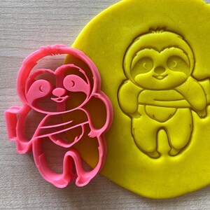May include: Pink plastic cookie cutter shaped like a sloth holding a cup of coffee. The cutter is next to a yellow piece of modeling clay with an impression of the sloth.