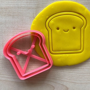 Può includere: Un tagliapasta di plastica rosa a forma di fetta di pane con un viso sorridente. Il tagliapasta è su una superficie gialla.